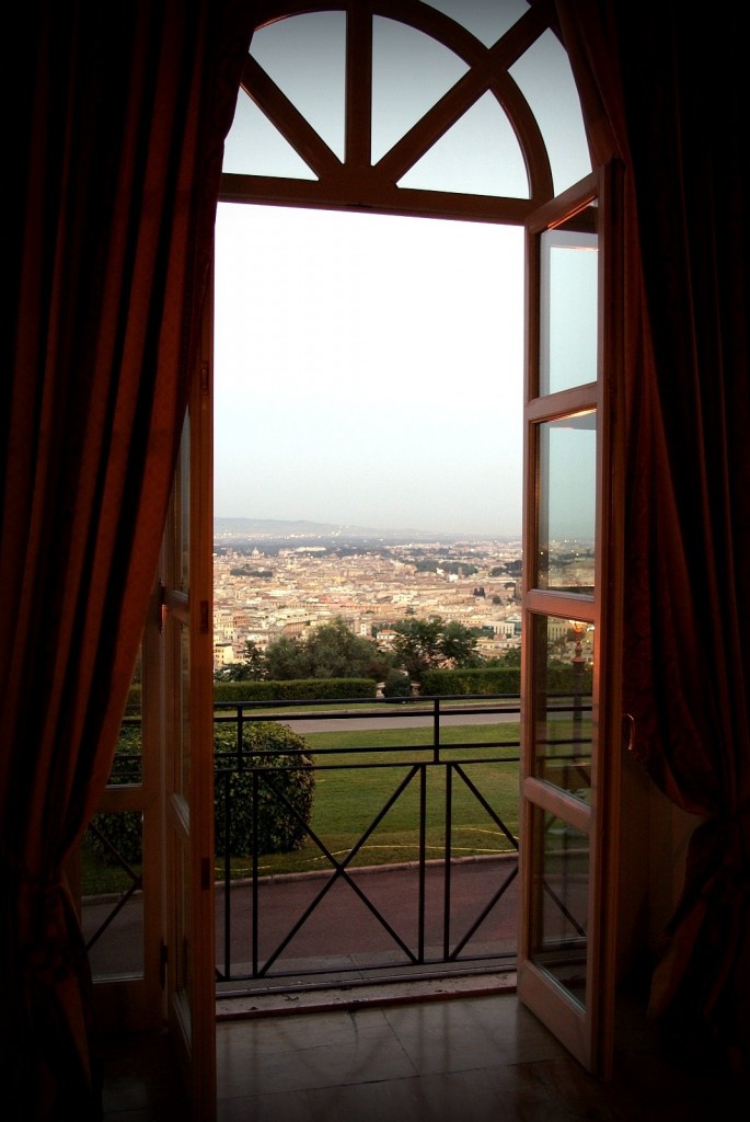 A beautiful tall window with a curved top, framed with red curtains looks out past a verandah and a lawn to city buildings in the distance.