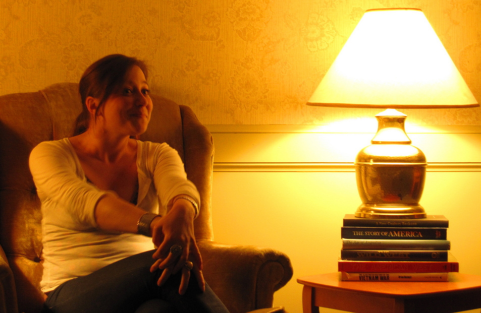 learn from personal stories: a photograph of a woman storyteller sitting in an armchair with a lamp on top of a pile of books on a table next to her
