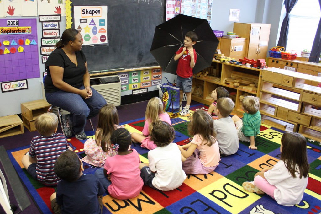 Do beats Show and Tell: a photograph of a kindergarten class with children sitting on the floor and a teacher and a child with an umbrella up front. Photo attribution: Flickr user wwworks.