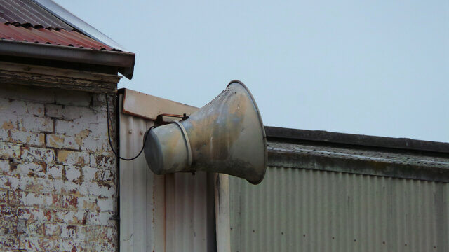 a photograph of a large rusty loudspeaker mounted on a corrugated metal wall. Photo attribution: Flickr user theenmoy
