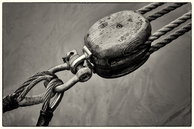 A conference connection economy: A black and white photograph of a wooden pulley. Two sisal ropes loop around the pulley on its right. On the left of the pulley is a metal shackle with two metal ropes attached to it.