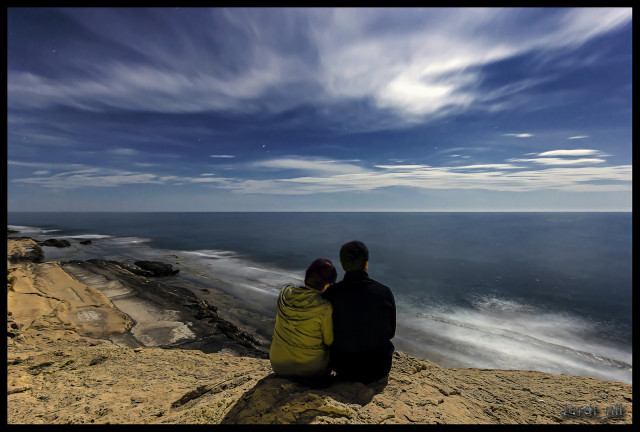 passion and engagement: photograph of the backs of a happy couple, arm-in-arm, sitting on the shoreline looking out to sea. Photo attribution: Flickr user 98810885@N07