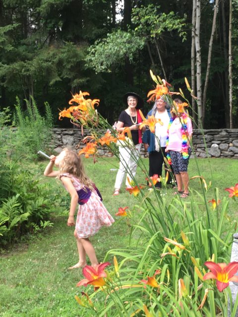 facilitate connection: A photograph. In a grassy cemetery with daylilies in the foreground and a stone wall and trees in the background, a child blows a horn while three smiling women look on.