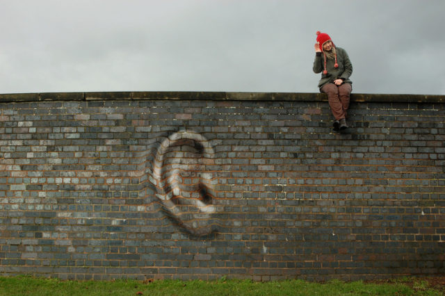 Conferences That Work meeting design: A photograph of a person sitting on top of a high brick wall with their hand cupped around their right ear. The wall displays an ear distorted out of the brick surface.