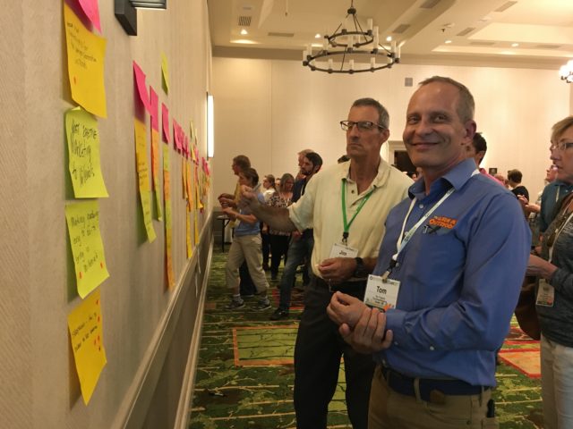 A photograph of a smiling conference participant who, with others, is dot voting on session topics written on large sticky notes posted on a wall