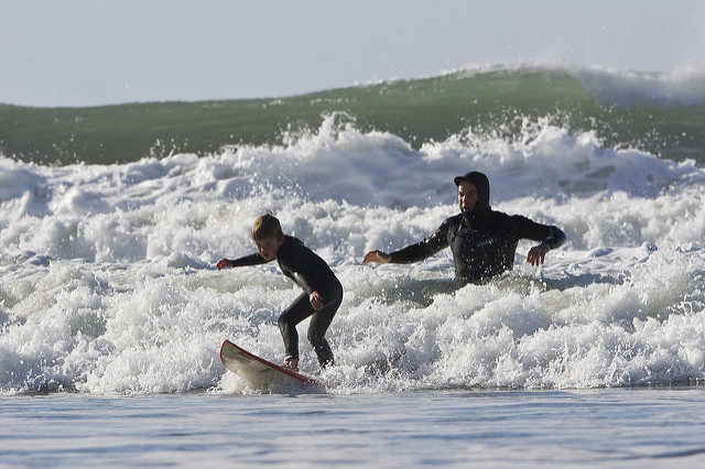 experiential learning: photograph of a young boy learning to surf in the ocean with his father standing just behind him. Photo attribution: Flickr user mikebaird