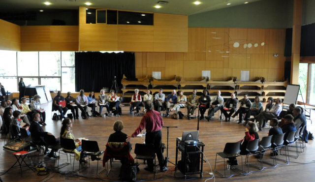 The best way to fundamentally improve your dull conference: Photograph of Adrian Segar [back to the camera, purple shirt] facilitating at a Conference That Work. Participants are sitting in a single large circle in a large wood-paneled hall.