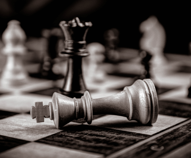 The declining influence of leadership positional power in a network society: A black and white closeup photograph of the corner of a chessboard. A white King piece is on its side, in front of a black Queen.