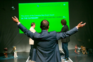 innovative conference competition: photograph of two conference participants facing each other in front of a projected award screen. In the foreground, an emcee stands with his back to the camera and his arms raised upwards.