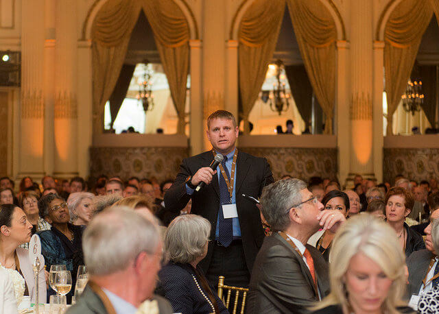 a meeting question that isn't: photograph of a man wearing a name badge standing in the middle of a seated audience talking into a microphone