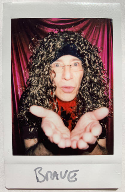 becoming brave: an instant photograph of Adrian Segar wearing a wig and makeup, holding out his hands towards the camera, with a red curtain behind him. The photo is labeled in marker "BRAVE".