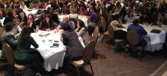 improve your facilitation: a photograph of a Solution Room conference session, with participants talking animatedly in groups of eight at round tables