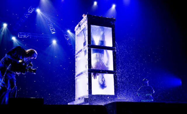 the second sort of escapologist: a photograph of a man upside down in a closed vertical glass case full of water. Image: Andrew Basso, The Escapologist. Photo Credit: Joan Marcus