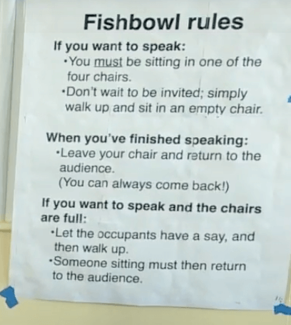 A photograph of a poster displaying Fishbowl Rules If you want to speak: —You must be sitting in one of the four chairs. —Don’t wait to be invited; simply walk up and sit in an empty chair. When you’ve finished speaking: —Leave your chair and return to the audience. (You can always come back!) If you want to speak and the chairs are full: —Let the occupants have a say, and walk up. —Someone sitting must then return to the audience.