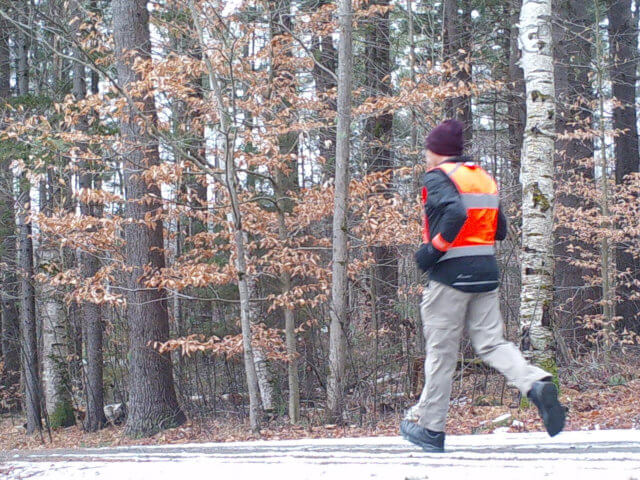 The author, Adrian Segar, running in winter gear in the Vermont woods. There's a little snow and ice on the gravel road.