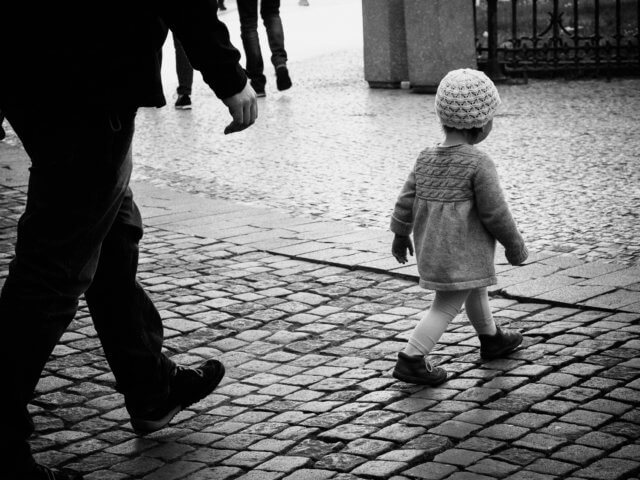 achieve success with one small step: a black-and-white photograph of a small child walking away from the camera on a brick-paved street. The child is followed by an adult whose step mirrors the child's.