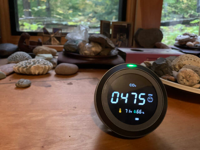 Photograph of an air quality tool meter laying on a desk with rocks and corals in the background. The cylindrical meter is showing a CO2 reading of 475 parts per million, a temperature of 71°F, and 66% humidity.