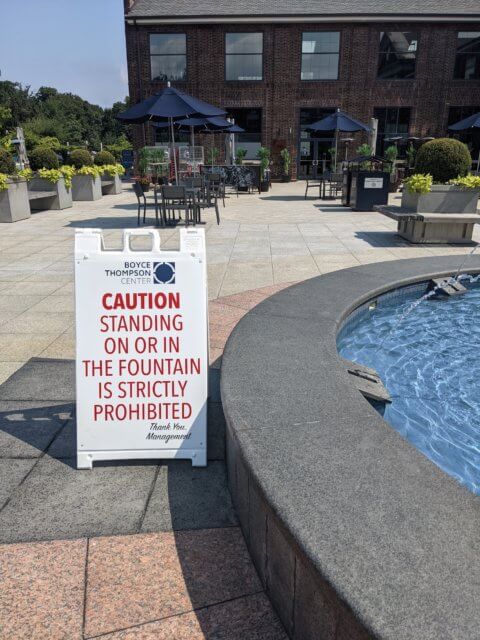 Make the meeting bigger! Photograph of an outdoor circular fountain with a low, wide grey wall around it. A white sign next to the fountain says, in red letters, "CAUTION STANDING ON OR IN THE FOUNTAIN IS STRICTLY PROHIBITED. Thank you. Management."