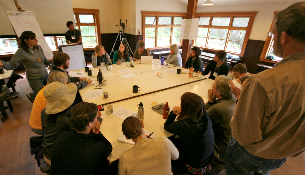 Photograph of a small group of people in a sunlit room. Three people are standing, the rest sit gathered around a set of tables pushed together. Several flipcharts are full of writing. People are talking, listening, and taking notes.